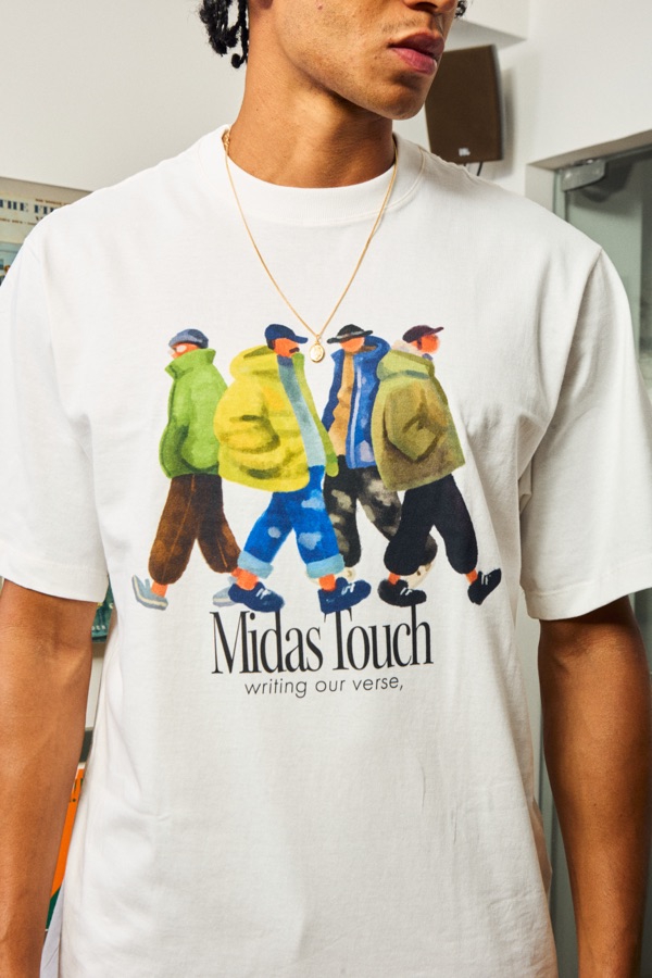 Camiseta branca estampada com ilustração colorida de pessoas caminhando e o texto “Midas Touch, writing our verse”